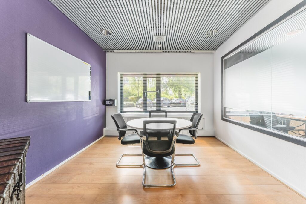 Vergaderruimte met ronde tafel, vijf stoelen, paarse muur met whiteboard en uitzicht op parkeerplaats aan de Prof. W.H. Keesomlaan.